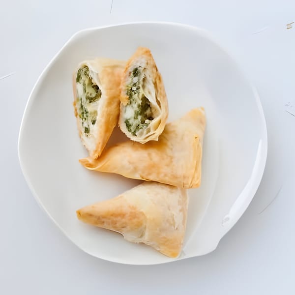 Three golden-brown spanakopita pastries, one cut open to reveal spinach and feta filling, on a white plate.