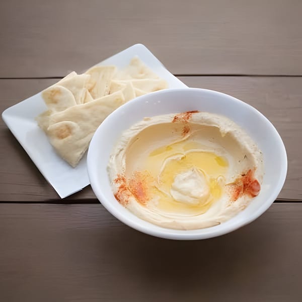 A white bowl of hummus drizzled with olive oil and paprika, with pita bread on a plate.